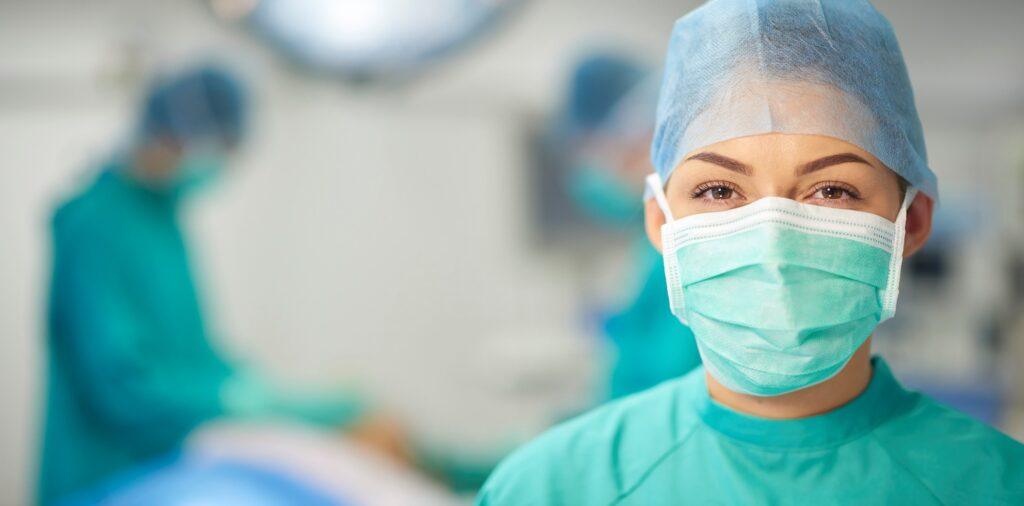 a young theatre nurse wearing all the appropriate surgical wear is looking to camera and smiling. In the background two surgeons can be seen working on a patient on the operating table